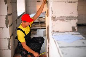 bauarbeiter auf der baustelle misst die länge der fensteröffnung und der mauer mit einem maßband. Häuschen bestehen aus porösen Betonblöcken, Arbeitskleidung - Overall und Baseballkappe foto