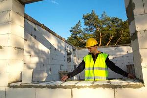 Bauarbeiter auf der Baustelle misst die Länge der Fensteröffnung und der Wand mit Maßband. Cottage bestehen aus Porenbetonblöcken, Schutzkleidung - Helm und Weste foto