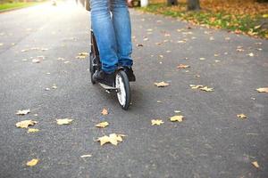 Die Beine eines Mannes in Jeans und Turnschuhen auf einem Roller im Park im Herbst mit gefallenen trockenen gelben Blättern auf dem Asphalt. Herbstspaziergänge, aktiver Lebensstil, umweltfreundlicher Transport, Verkehr foto