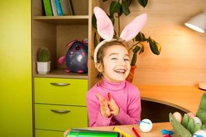 ein mädchen in den ohren eines osterhasen malt eier mit einem filzstift im wohninnenraum. Kunsthandwerk, Vorbereitung auf einen religiösen Feiertag, ein Tablett mit Eiern, Hasenohren aus Plastilin foto