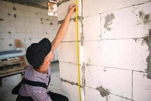 bauarbeiter auf der baustelle misst die länge der fensteröffnung und der mauer mit einem maßband. Häuschen bestehen aus porösen Betonblöcken, Arbeitskleidung - Overall und Baseballkappe foto