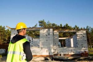 projektarchitekt steht in der nähe der baustelle eines hauses, dessen wände aus einem porösen betonblock und einem ziegel bestehen. Bauarbeiter mit gelbem Schutzhelm und Warnweste. foto