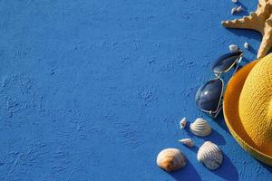 Rahmen mit Strandzubehör zu einem nautischen Thema Gelber Strohhut, Sonnenbrille, Seesterne und Muscheln auf blauem Hintergrund. Urlaubskonzept, Seereise, UV-Schutz, Schwimmen. Platz kopieren. flachgelegt foto