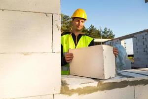 Der Baumeister hält einen Porenbetonblock in den Händen - das Mauerwerk der Hauswände. Bauarbeiter in Schutzkleidung – Helm und Warnweste. foto