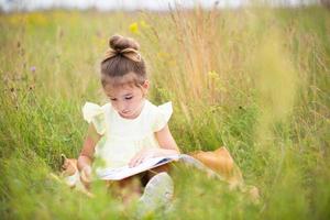 Mädchen in einem gelben Kleid sitzt im Gras auf einer Decke auf einem Feld und liest ein Buch aus Papier. Internationaler Kindertag. Sommerzeit, Kindheit, Bildung und Unterhaltung, Hüttenkern. Platz kopieren foto