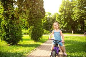 ein mädchen in einem jeansoverall fährt im sommer in einem grünen park ein lila laufrad. aktive Unterhaltung für Kinder, Roller für Kleinkinder, glückliches Baby. Platz kopieren foto