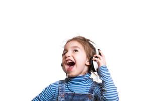 Kleines Mädchen in einem gestreiften blauen Rollkragenpullover hört Musik mit weißen Kopfhörern und singt laut mit offenem Mund. Spaß, Geschrei, Kindergefühle. weißer hintergrund, isolieren, kopierraum foto