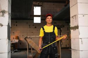 bauarbeiteringenieur auf der baustelle misst die länge der fensteröffnung und der ziegelwand mit einem maßband. Mann in Arbeitskleidung - Overall und Mütze foto