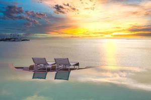 sommerlicher Liebes- und Romantiktisch für ein romantisches Abendessen mit Infinity-Pool-Reflektionsstühlen unter Sonnenuntergang und Meer im Hintergrund. Luxus-Dinner, romantisches Abendessen für Paare foto