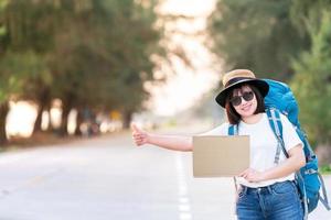 Fröhlicher lächelnder Anhalter asiatischer Frauenrucksack, der am Wochenende auf den Autotransport wartet, um bei Sonnenuntergang im Urlaub zu reisen und zu reisen foto