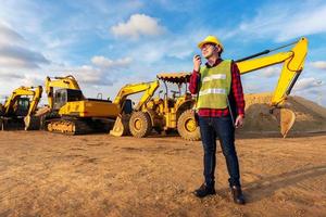 asiatische techniker bauingenieur sprechen mit team per walkie-talkie halten laptop-computer oder tablet für transportbauarbeiten mit bagger auf dem hintergrund der baustelle foto