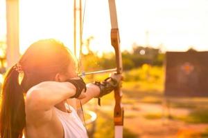 junge bogenschützin, bogenschießen, schießen mit bogen im naturfeld zum ziel, erfolgskonzept, auf dem feld für sportübungen bei sonnenuntergang foto