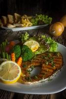 Lachssteak mit Soße auf einem weißen Teller mit Zitrone auf dem Teller und knusprigen Pommes Frites. Viele Gemüsesorten werden um die Schüssel herum auf dem Holzboden platziert. foto