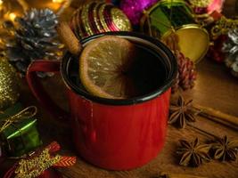 heißes getränk alkohol, roter glühwein in roten rustikalen bechern gewürzen zimtstange und zitrusfrüchten. dekorierte tanne festlich auf holztisch. feier weihnachten und frohes neues jahr konzept. foto