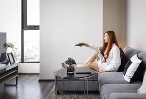 asiatische frau, die fernbedienung verwendet, um den kanal zu wechseln, während sie am wochenende entspannt zu hause auf dem bequemen sofa fernsehen foto