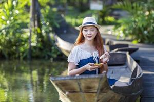 junge asiatische touristin reist mit einem holzboot auf dem schwimmenden markt in thailand und hat lokales straßenessen für das südostasiatische tourismuskonzept foto