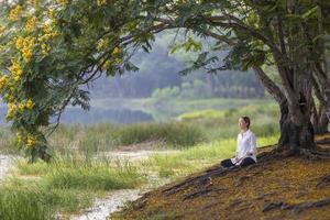 Frau, die entspannend Meditation im öffentlichen Park praktiziert, um im Sommer Glück aus innerer Friedensweisheit unter gelbem Blumenblütenbaum zu erlangen foto