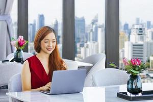Der luxuriöse asiatische Millionärs-CEO im roten Kleid arbeitet mit einem Laptop-Computer im prestigeträchtigen Büro mit Wolkenkratzerblick für ein wohlhabendes und gehobenes Lifestyle-Konzept
