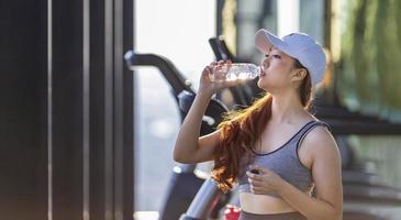 asiatische frau genießt die morgendliche übung im fitnessstudio, während sie eine pause macht, um mineralwasser zur erfrischung und rehydrierung zu trinken foto