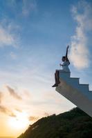 asiatische frau hebt die arme hoch und sitzt oben auf einer holztreppe mit sonnenuntergangshimmel im hintergrund foto