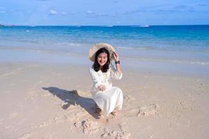 Attraktive asiatische Frau im weißen Kleid genießt es, am wunderschönen Inselstrand zu sitzen und Sand zu spielen foto