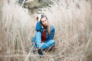 hübsches Mädchen sitzt in einem schönen Herbstgras. blue jeanyoung schönes Mädchen mit einem Lächeln in Jeanskleidung. Jacke. foto