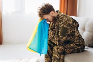 porträt eines emotionalen jungen ukrainischen patriotensoldaten in militäruniform, der im büro auf der couch sitzt und eine gelb-blaue flagge hält. foto