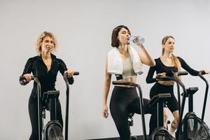 Junge Frauen, die Wasser trinken und Airbike für Cardio-Training im Cross-Training-Fitnessstudio verwenden foto