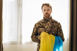 porträt eines emotionalen jungen bärtigen ukrainischen patriotensoldaten in militäruniform, der eine flagge im büro hält foto