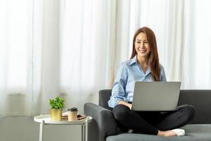 Fröhliche, lässige, schöne asiatische Frau, die an einem Laptop arbeitet, der als Freiberufler auf dem Sofa sitzt, Arbeit von zu Hause aus. foto