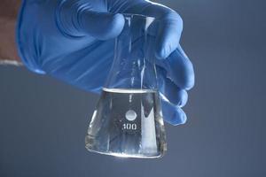 Eine Hand in einem medizinischen Handschuh hält eine Laborflasche mit Flüssigkeit auf grauem Hintergrund. das Konzept eines chemischen Labors. foto