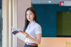 Schöne junge asiatische Studentin mit schwarzen langen Haaren in Schuluniform liest ein Buch in der Universitätsbibliothek. foto