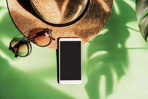 reisezubehör und leeres bildschirm-smartphone mit schatten von tropischem grün verlassen auf farbigem hintergrund mit kopienraum, sommerferienkonzept foto