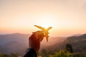 hand, die ein miniaturflugzeug mit bergblick bei sonnenaufgang, sommerferien und reisekonzept hält foto