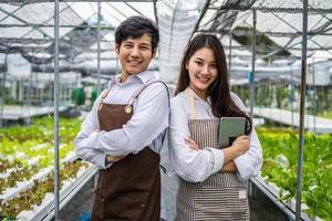 Neue Generation von lächelnden zwei jungen asiatischen Bauernpaaren, die in Gemüse-Hydrokulturfarmen arbeiten, erfolgreiche Besitzer von Hydroponik-Gemüsegärten, die in Gewächshausplantagen für gesunde Ernährung stehen foto