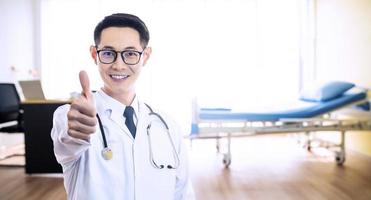 asiatischer guter arzt mann lächelt und zeigt daumen hoch für den besten gesundheits- und versicherungspaketplan für werbung, über dem bett im krankenhaushintergrund, kopierraum foto