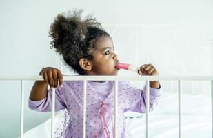 Afroamerikanisches süßes Babymädchen, das Eis auf dem Bett im Schlafzimmer isst foto