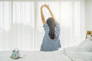 Rückansicht einer schönen jungen Frau, die morgens auf ihrem Bett sitzt und sich mit erhobenen Armen streckt, es gibt einen Wecker, der die Zeit angibt, gesunder Lebensstil, Wellness-Konzept, Freiraum foto