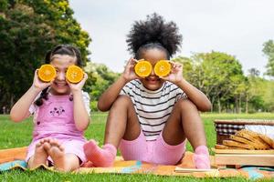 Zwei afroamerikanische kleine Mädchen sitzen auf der Matte und essen Orangenfrüchte im Herbstpark, spielen mit Orangenfrüchten foto