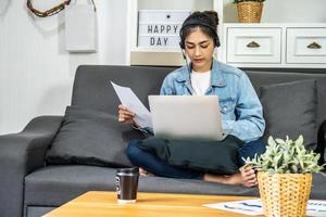 asiatische junge geschäftsfrau sitzt auf dem sofa in ihrem eigenen haus trägt kopfhörer videoanrufkonferenz mit laptop-computer mit stapel von diagramm- und diagrammpapier im büro zu hause, arbeit von zu hause aus foto