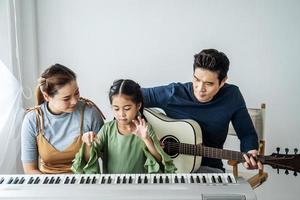 glückliche kleine asiatische tochter spielt klavier mit mutter und vater spielen zu hause gitarre, mutter bringt tochter das klavierspielen bei, sie spielen und singen lieder. Sie haben Spaß. foto