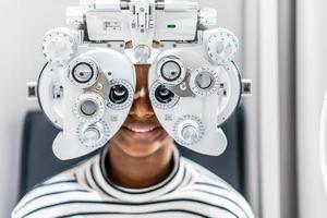 Lächelnde junge Frau afroamerikanische Afro-Haare, die einen Sehtest auf einem optischen Phoropter durchführen und ihr Auge mit einem Optometriegerät überprüfen foto