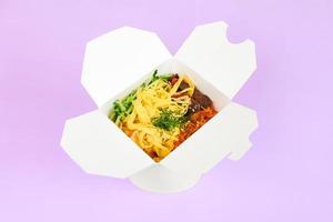 Reiswok mit Meeresfrüchten und Gemüse in weißer Box isoliert auf violettem Hintergrund, Fast-Food-Lieferung foto