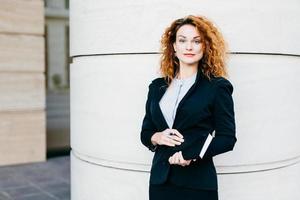 geschäftsfrau mit lockigem hellem haar, rot bemalten lippen, trägt weiße bluse, schwarze jacke und rock, hält notizbuch mit stift in der hand und schreibt notwendige notizen. menschen, erfolg und geschäftskonzept foto