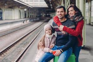 Foto einer freundlichen Familie, gute Beziehung, Reise im Urlaub, Pose am Bahnsteig des Bahnhofs. schöne frau umarmt ehemann, ihre kleine tochter steht in der nähe der eltern in der nähe des gepäcks