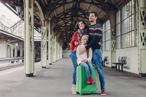 Zufriedene Frau und Mann kuscheln in Liebe, ihre Tochter sitzt am Koffer, posieren gemeinsam auf Bahnsteig, schauen in die Ferne, warten auf den Zug, haben erfreute Gesichtsausdrücke. glückliche familienreise ins ausland foto