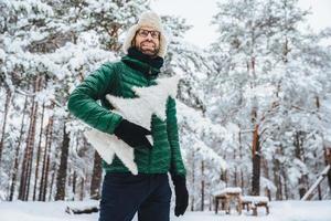 Fröhliches männliches Model in Winterkleidung, hält weißen künstlichen Tannenbaum, steht im Winterwald, hat ein angenehmes Lächeln, genießt schöne Landschaften. menschen, ferien, erholungskonzept foto