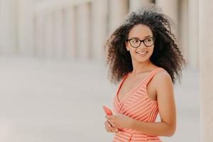 zufriedenes tausendjähriges Mädchen mit lockigem Haar, trägt rosa Kleid, optische Brille, konzentriert sich mit einem Lächeln zur Seite, steht im Freien, wartet auf Anruf, genießt Hochgeschwindigkeits-Internetverbindung, aktualisiert neue Anwendung foto