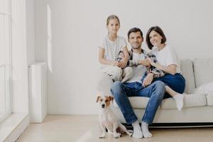 horizontale aufnahme einer liebevollen familie, die zusammen auf der couch in einem leeren, geräumigen raum mit weißen wänden posiert, ihr lieblingshund sitzt auf dem boden. Platz beiseite kopieren. glückliches weibliches Kind, froh, bei Mama und Papa zu sein foto