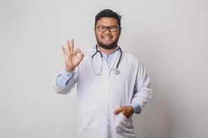 glücklicher männlicher Doktor, der ok lokalisiert auf weißem Hintergrund gestikuliert foto